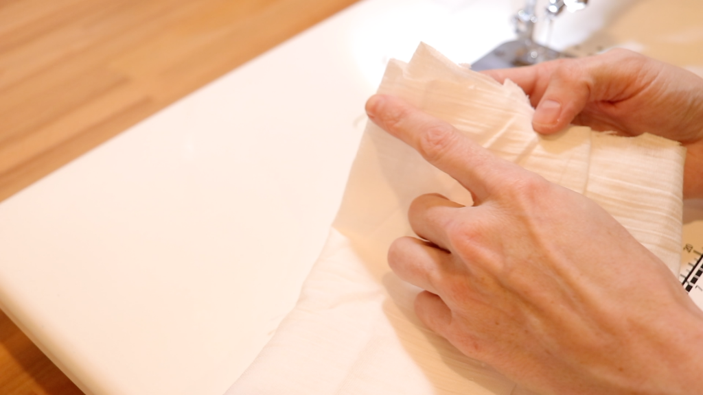 With a loose stitch sewn in the fabric, a womans hands are demonstrating how to gather the fabric by pulling on the white thread.