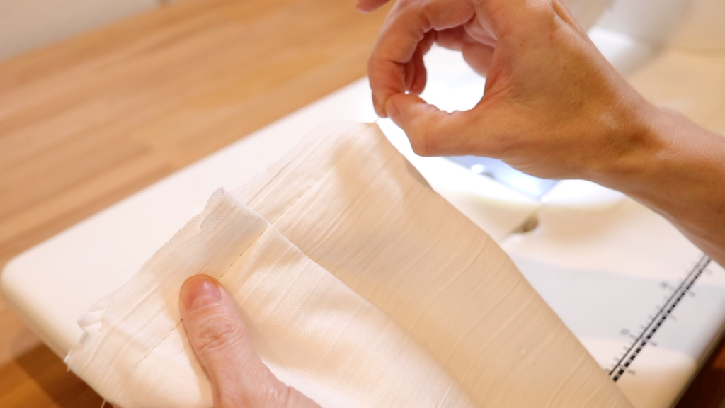 With a loose stitch sewn in the fabric, a womans hands are demonstrating how to gather the fabric by pulling on the white thread. 