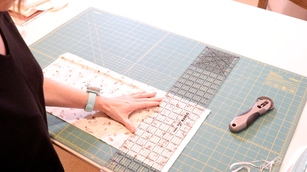 A womans hand is holding a ruler to cut the selvage edge of her large piece of fabric. A rotary cutter is sitting nearby. 