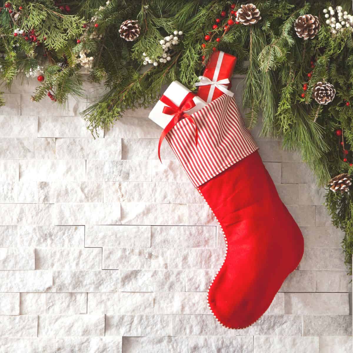 A beautiful red and white Christmas stocking hanging in front of a white brick fireplace. Green garland is strung above it with small bunches of red berries in the garland. Red and white Christmas packages peaking out of the top of the stocking.