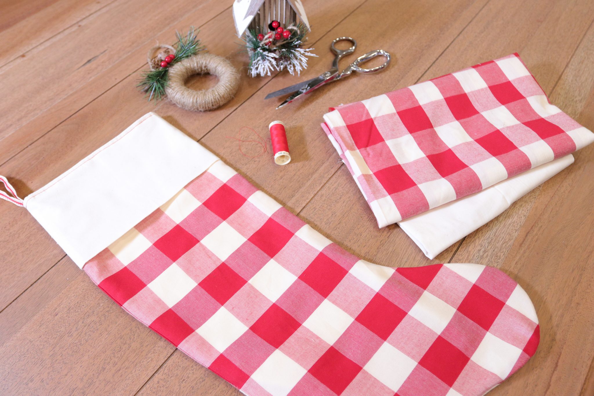 A bright red and white checked Christmas Stocking with a white cuff and hanging loop is sitting on a farmhouse table. The stocking is sitting next to a spool of red thread, a scrap pile of the fabric used, and a tiny Christmas birdhouse ornament.