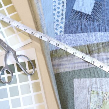 A newly finished blue and green log cabin quilt sitting on a table near a pair of silver scissors, a tape measure and a color chart resembling colors used in the quilt.