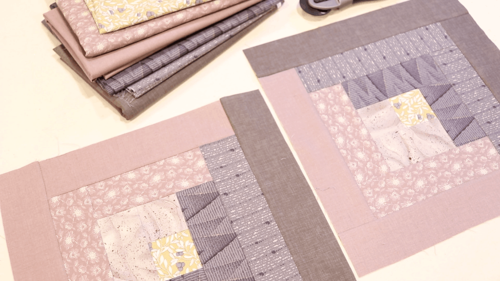 a table has 2 neatly pieced log cabin quilt blocks squares. The squares are made using light purple and pale pink fabrics. A small stack of fabric is nearby and a rotary cutter.