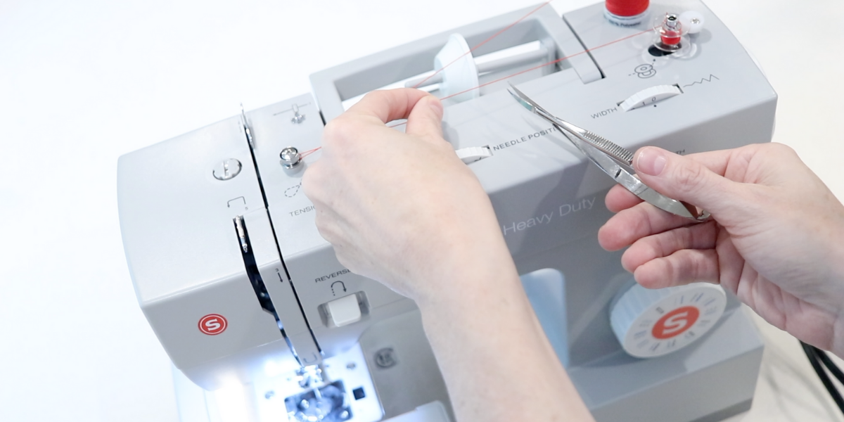 a woman's hand using small scissors to clip the red thread on a sewing machine. Blog post about how to thread a sewing machine