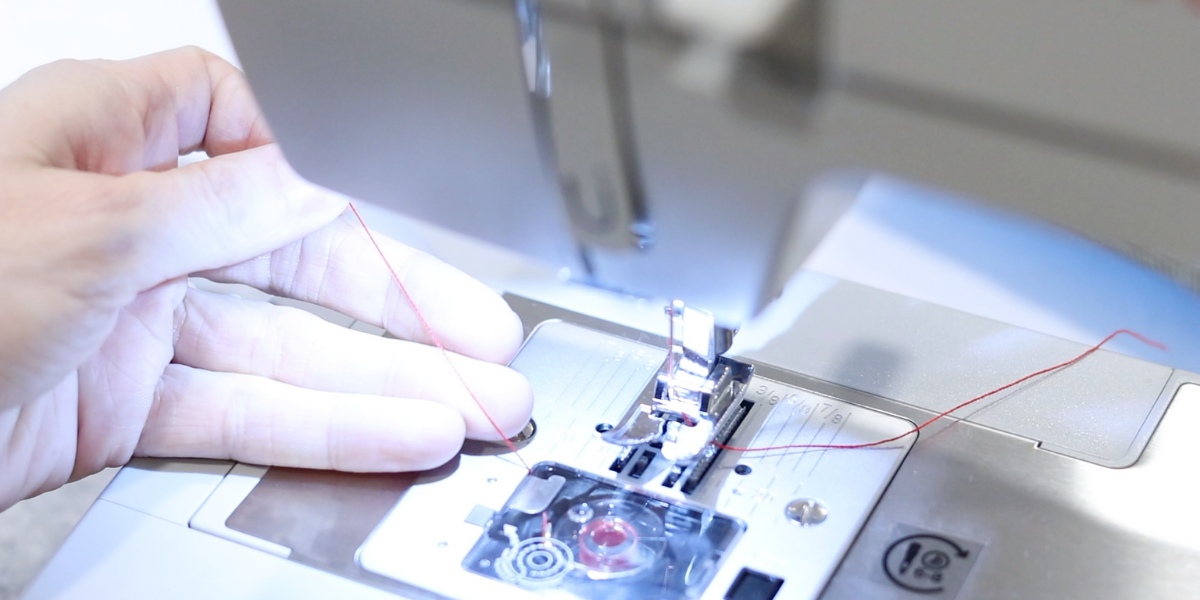 left hand holding a red thread tightly as it is passed around a sewing machine bobbin case.