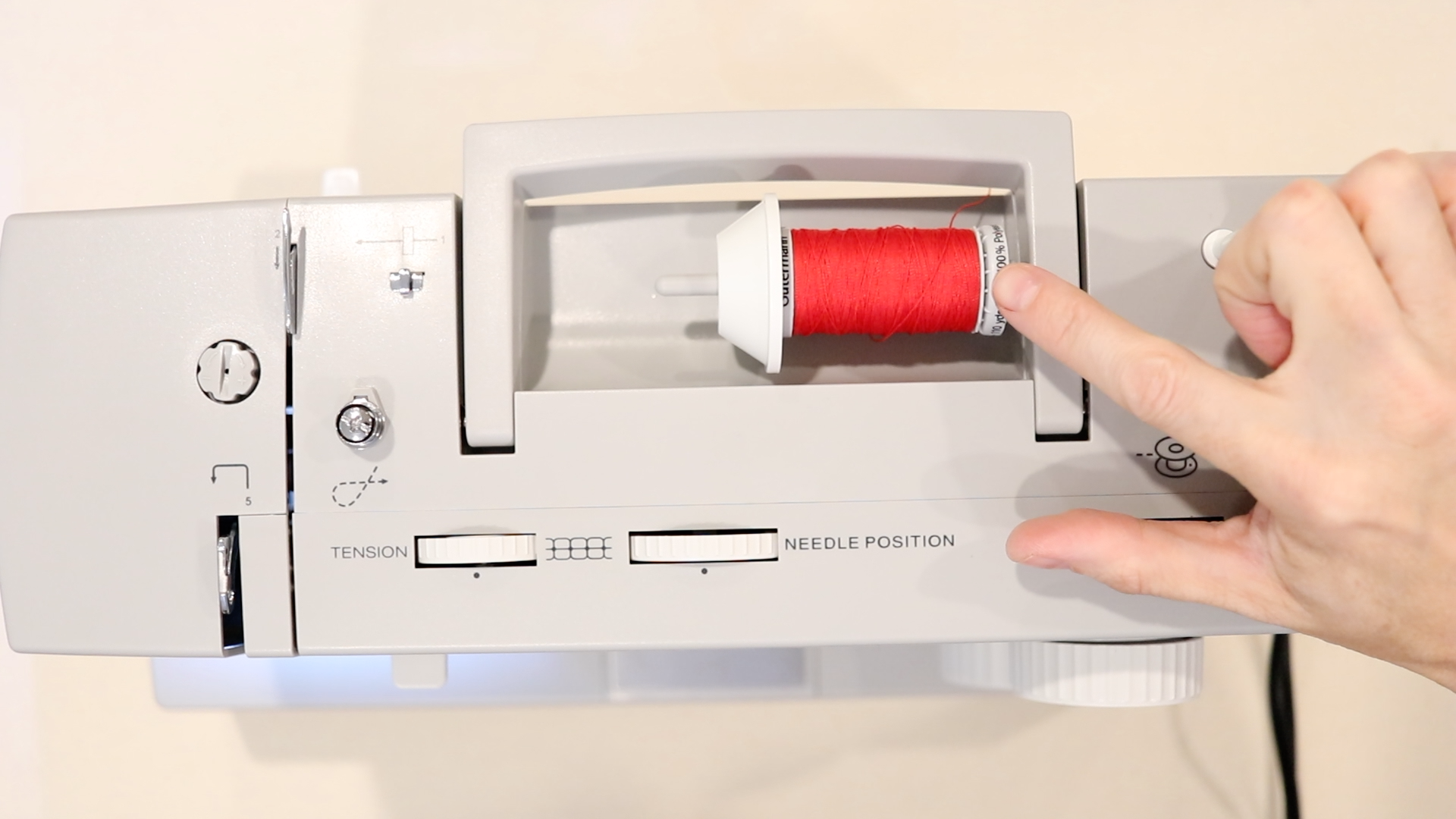 Hand pointing at a spool of bright red thread on the top of a gray sewing machine. A spool cap is in place to keep thread from coming off while sewing.