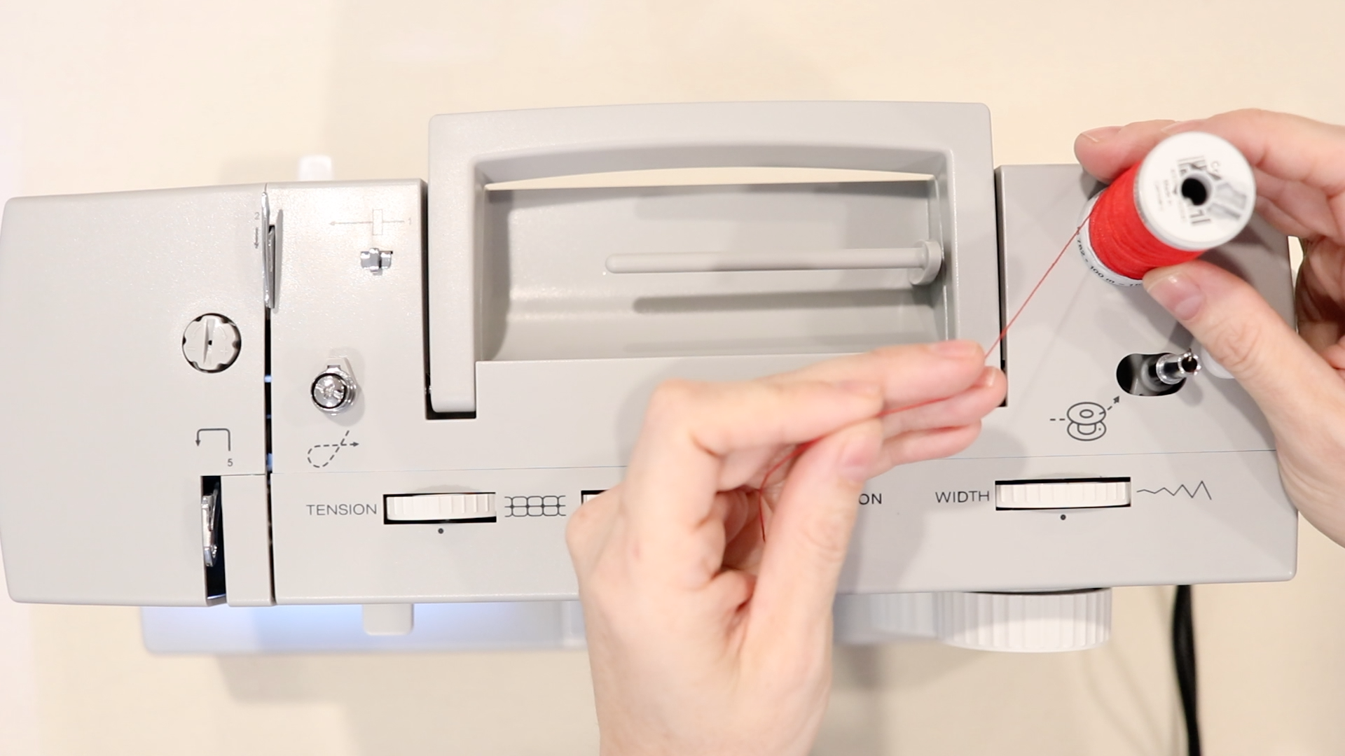 hands placing a red spool of thread on a vertical spool pin that's on a gray sewing machine.