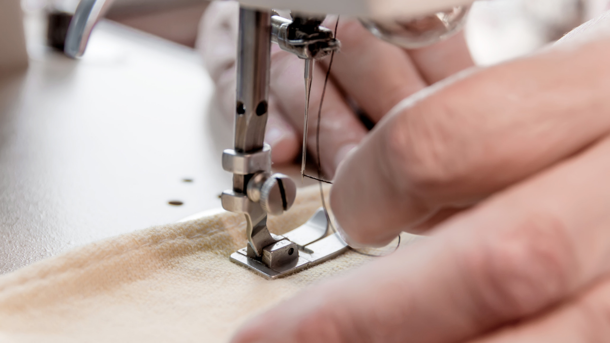 A hand threading a sewing machine needle from left to right through the eye of the sewing machine needle