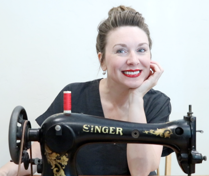 Brie is sitting behind a beautiful black vintage Singer treadle sewing machine. She is wearing a black shirt, her hair is neatly put into a bun and is wearing a bright red Chanel lipstick and smiling at the camera