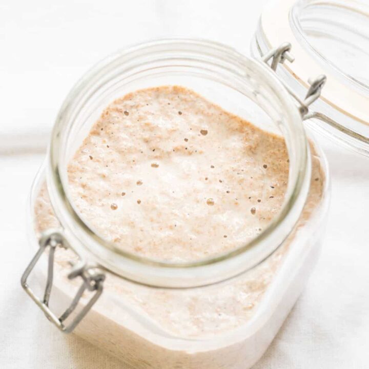 A clear, square jar with an open top filled with an active, bubbly sourdough starter.
