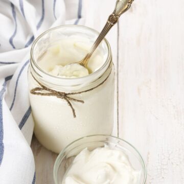 a canning jar full of white homemade sour cream. A piece of jute twine is tied around the top of the jar and a silver spoon is resting inside. A small white dish of sour cream resting nearby.
