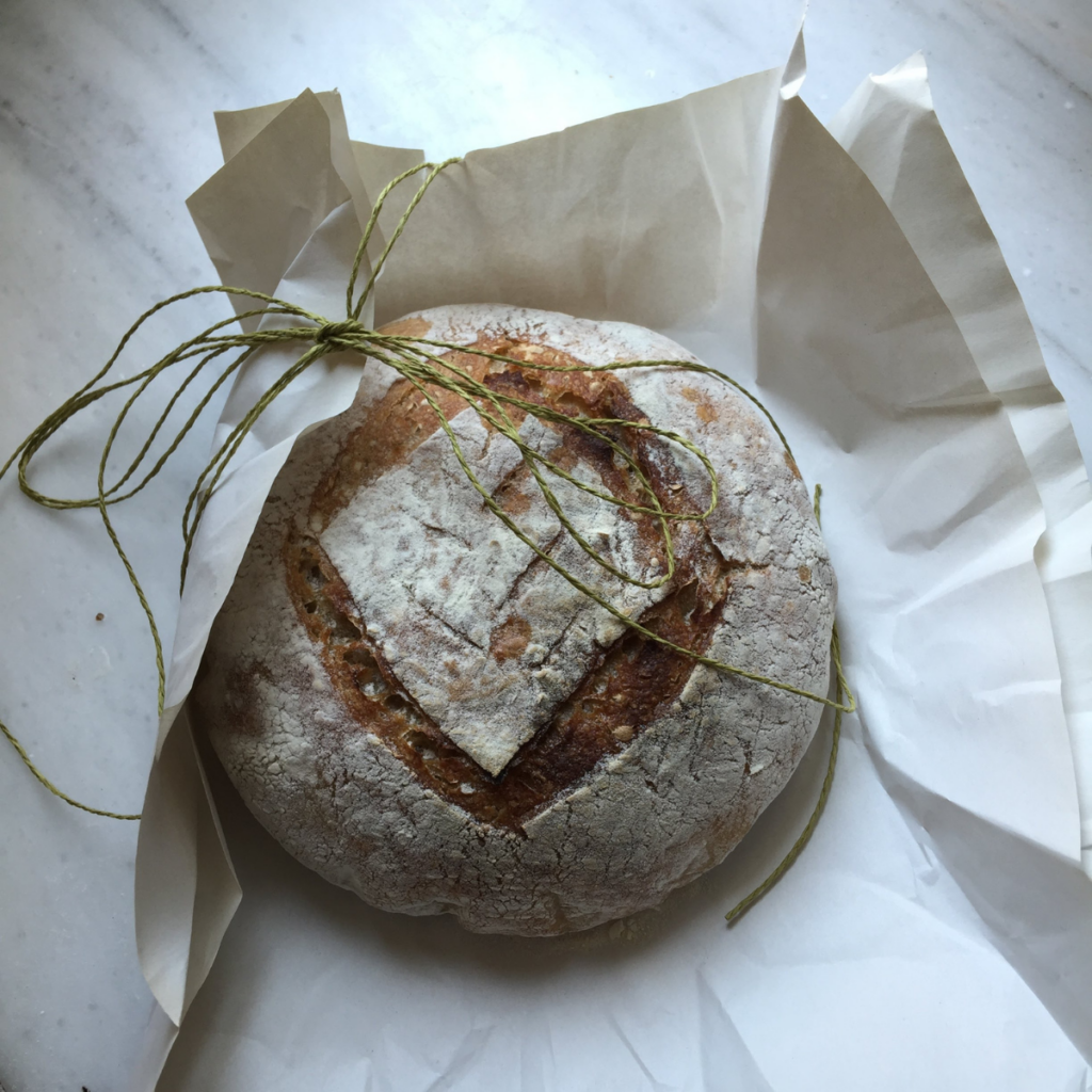 A fresh baked loaf of sourdough bread sitting on a piece of parchment paper. Loaf is being wrapped in paper with a piece of jute twine. 