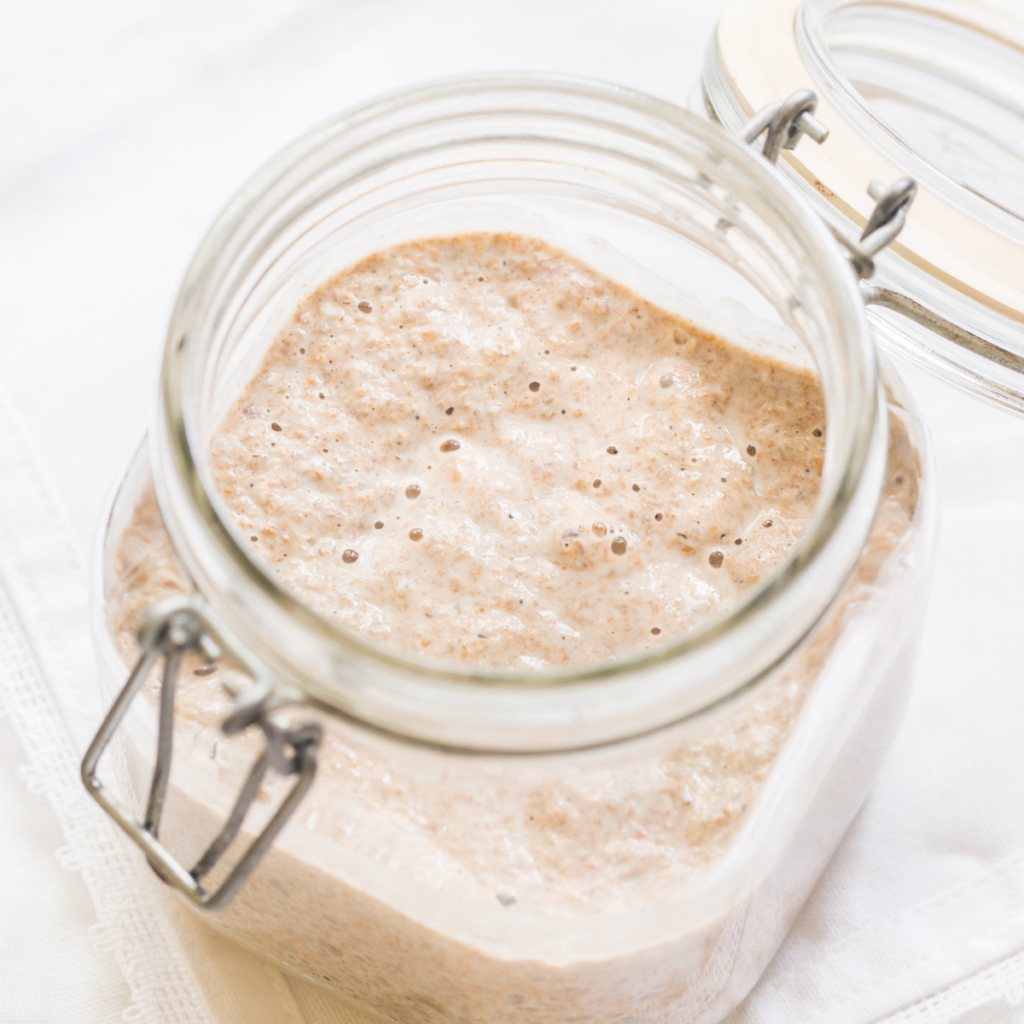 a clear glass jar with a bubbling, active sourdough starter inside. Jar is sitting on a white tea towel on a table top. 