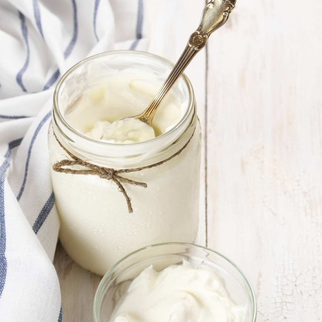 a homemade jar of white sour cream sitting on a  kitchen table with a white and blue striped towel behind it. An antique spoon resting inside the jar that has been tied with a piece of jute twine