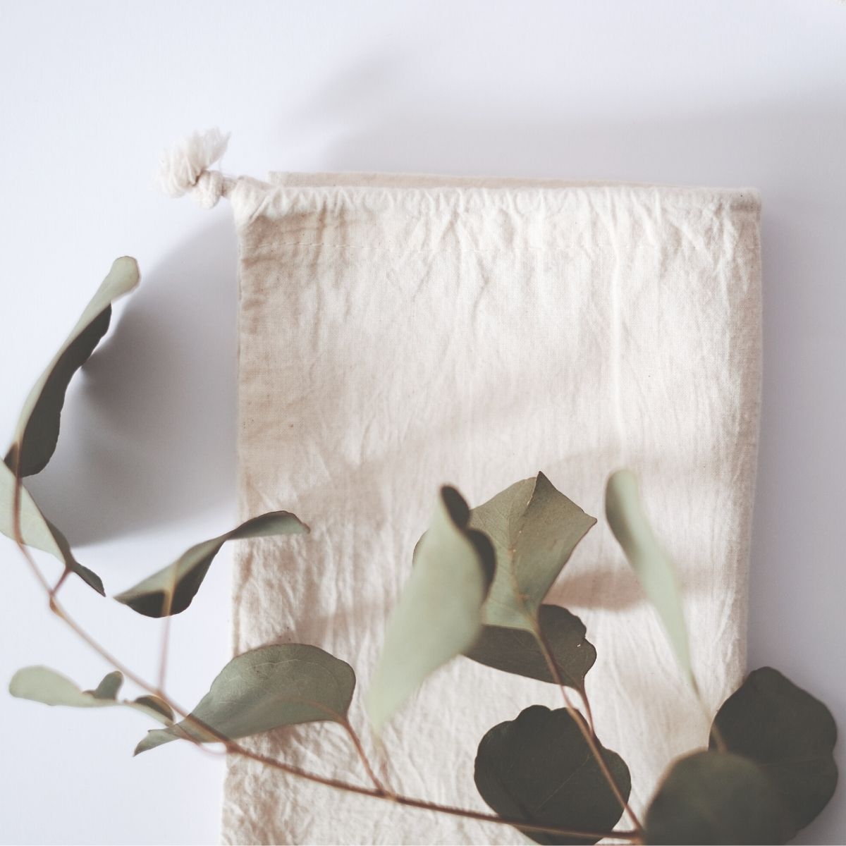 a small muslin drawstring bag sitting on a white table top. The muslin bag has a small branch of dried eucalyptus sitting onto of it. The sun is beaming in through a window from the left side.