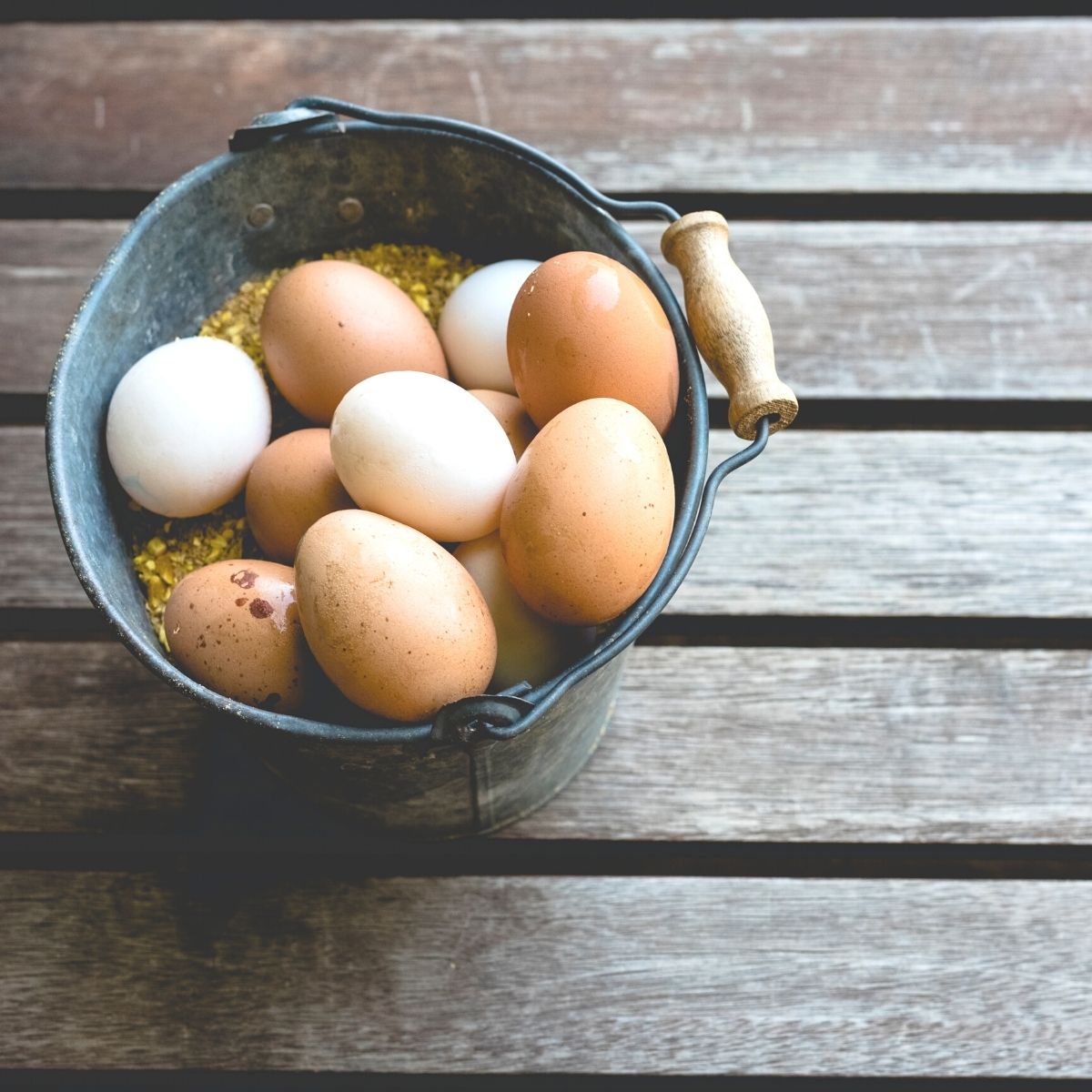 a bucket of freshly collected chicken eggs sitting on an old plank deck. The bucket contains a variety of white and brown eggs.