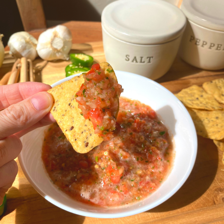 A fresh bowl of homemade salsa. A hand holding a tortilla chip that has salsa on it. All the ingredients are nicely placed on a beautiful teakwood cutting board. A salt and pepper pot is in the background.