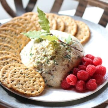 A half dome of savory cheese spread set on a crisp white plate. A generous portion of red raspberries and multigrain crackers sitting on the sides of it. A Green Bay leaf sprig rests on the top.