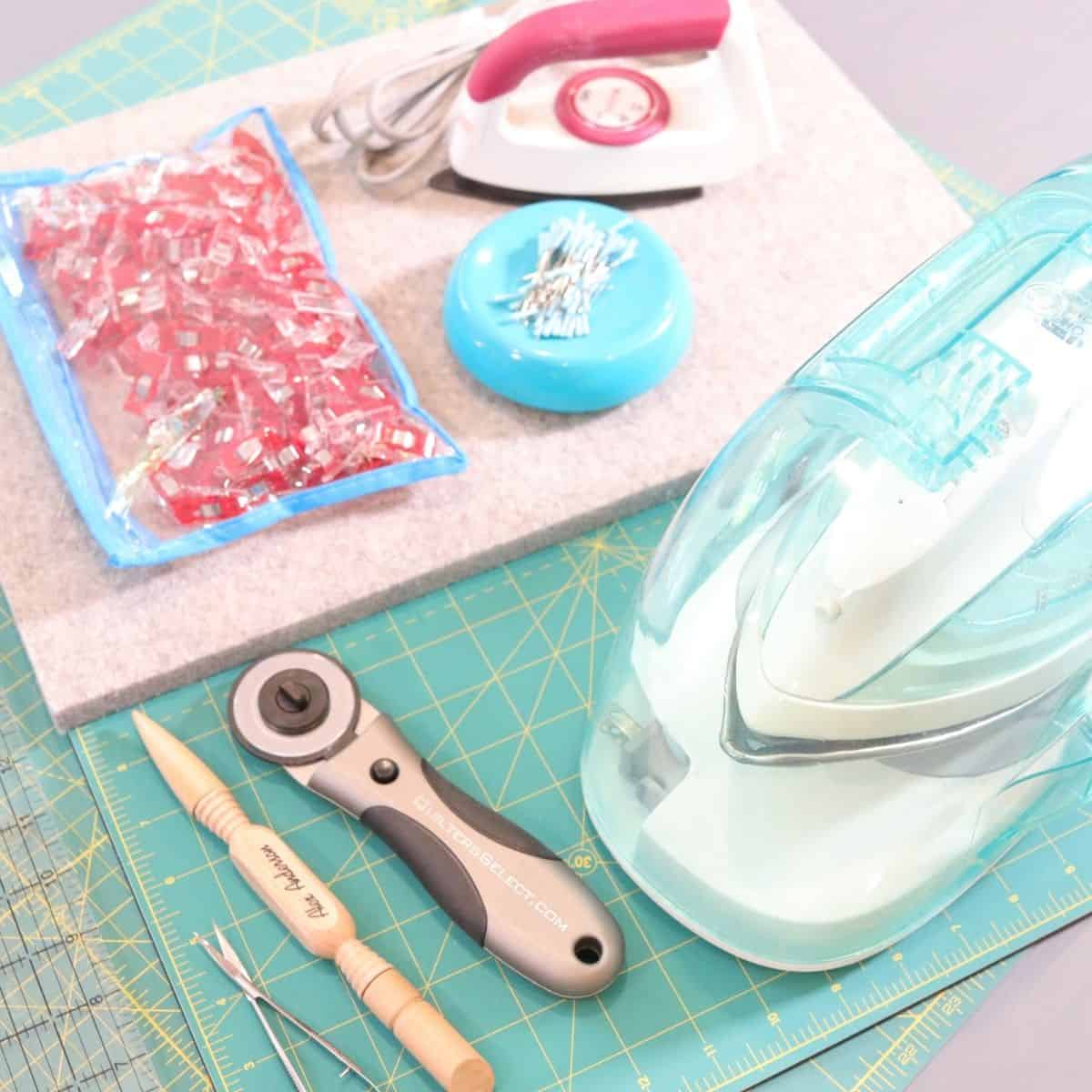 Sewing supplies gathered together on a cutting table. Sewing pins, an iron, a wooden 4 in 1 sewing tool and a metal rotary cutter.