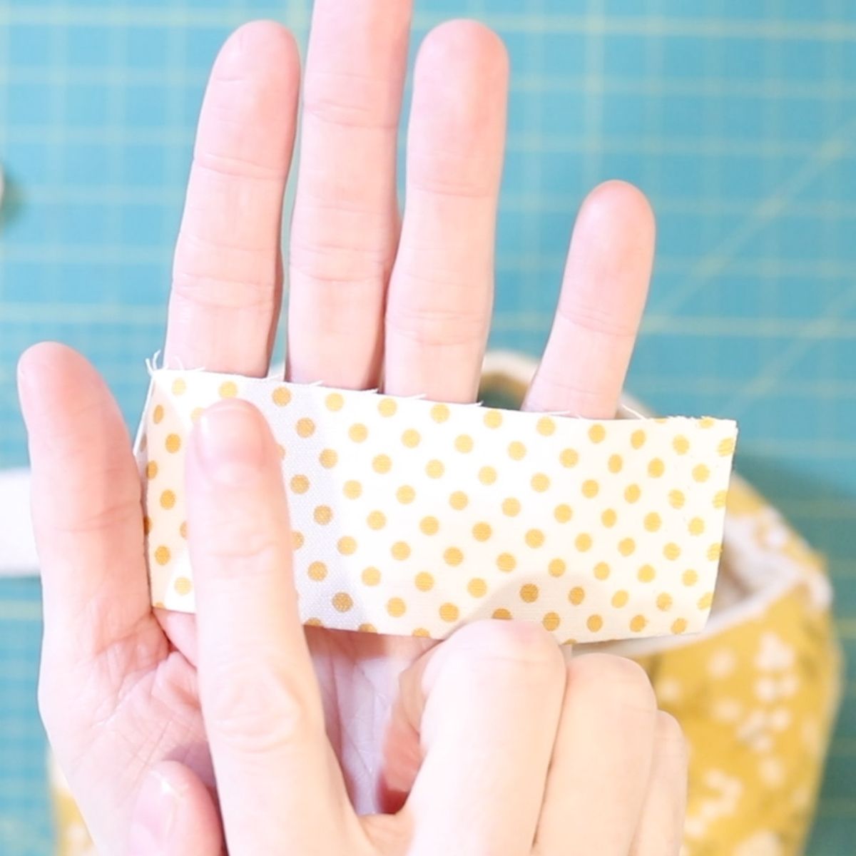 a handing holding a strip of yellow and white polka doted fabric binding pointing to the raw edge of the fabric