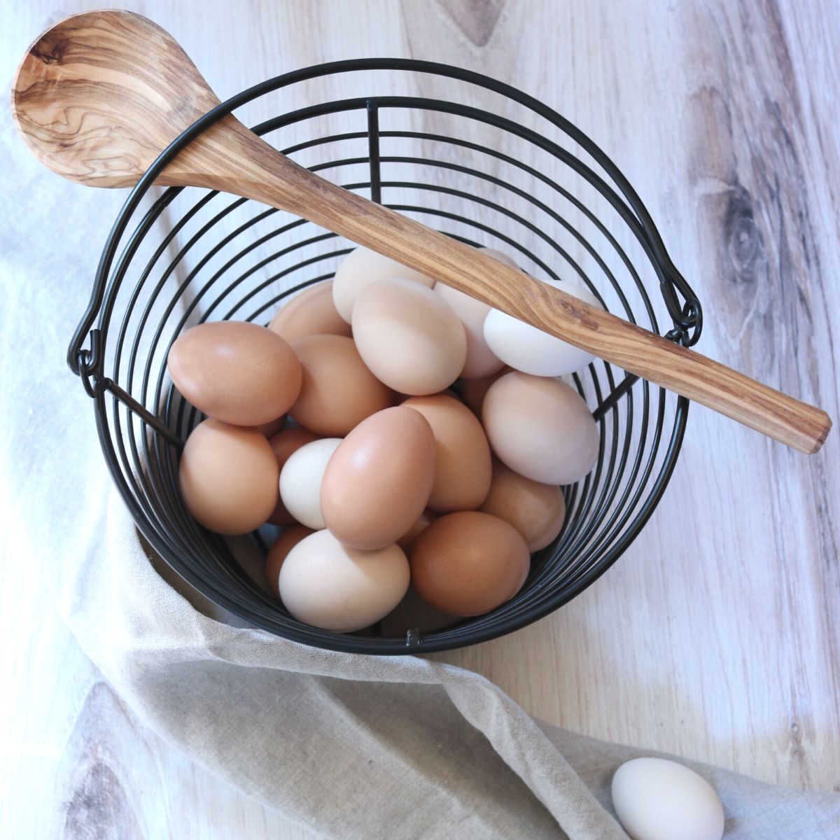 A black wire basket full of freshly collected chicken eggs. A wooden spoon rests on the top of the basket.