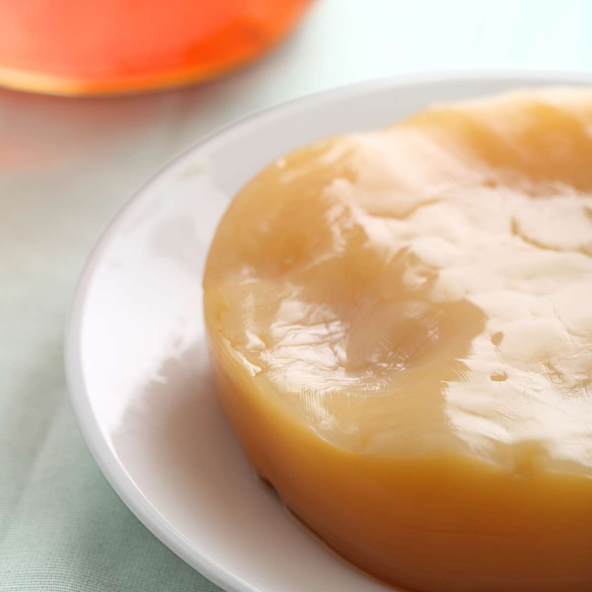 A close up picture of a healthy Kombucha SCOBY. It is tan in color and sitting on a white plate that is resting near a jar of kombucha tea.