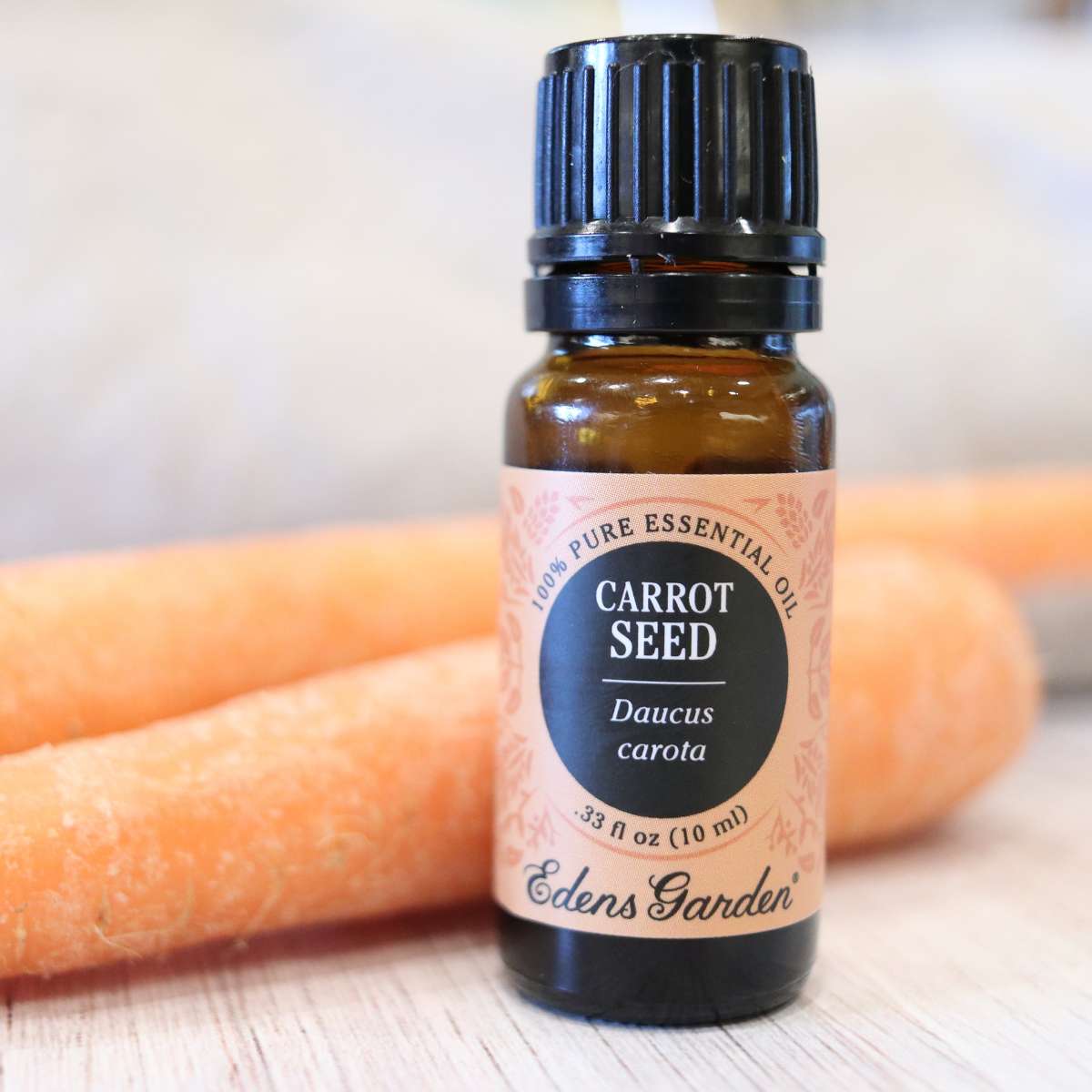 A bottle of carrot seed essential oil with a pale orange wrapper sitting on a table. Two brightly colored orange carrots can also be seen in the picture.