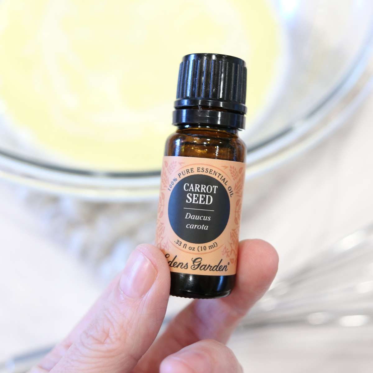 A bottle of carrot seed essential oil in a glass amber bottle with a black cap. A bowl of homemade DIY sunscreen is in the background.