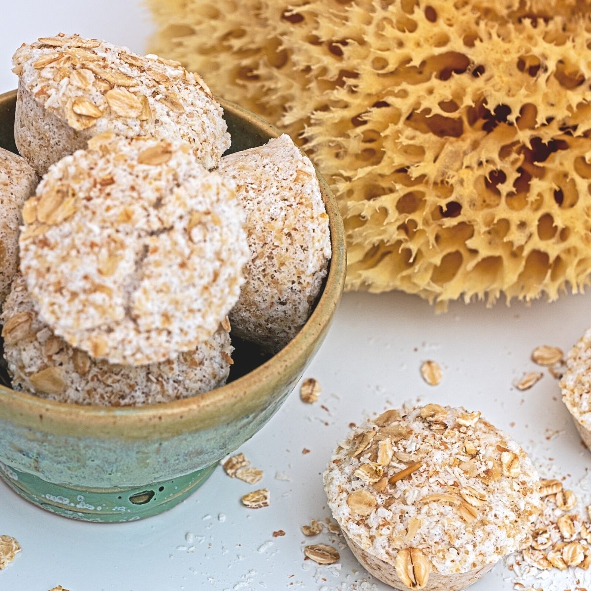 A beautiful seafood green bowl with DIY colloidal oatmeal bath soak. A yellow natural sea sponge rests behind the bowl. Pieces of oatmeal are scattered around the table.