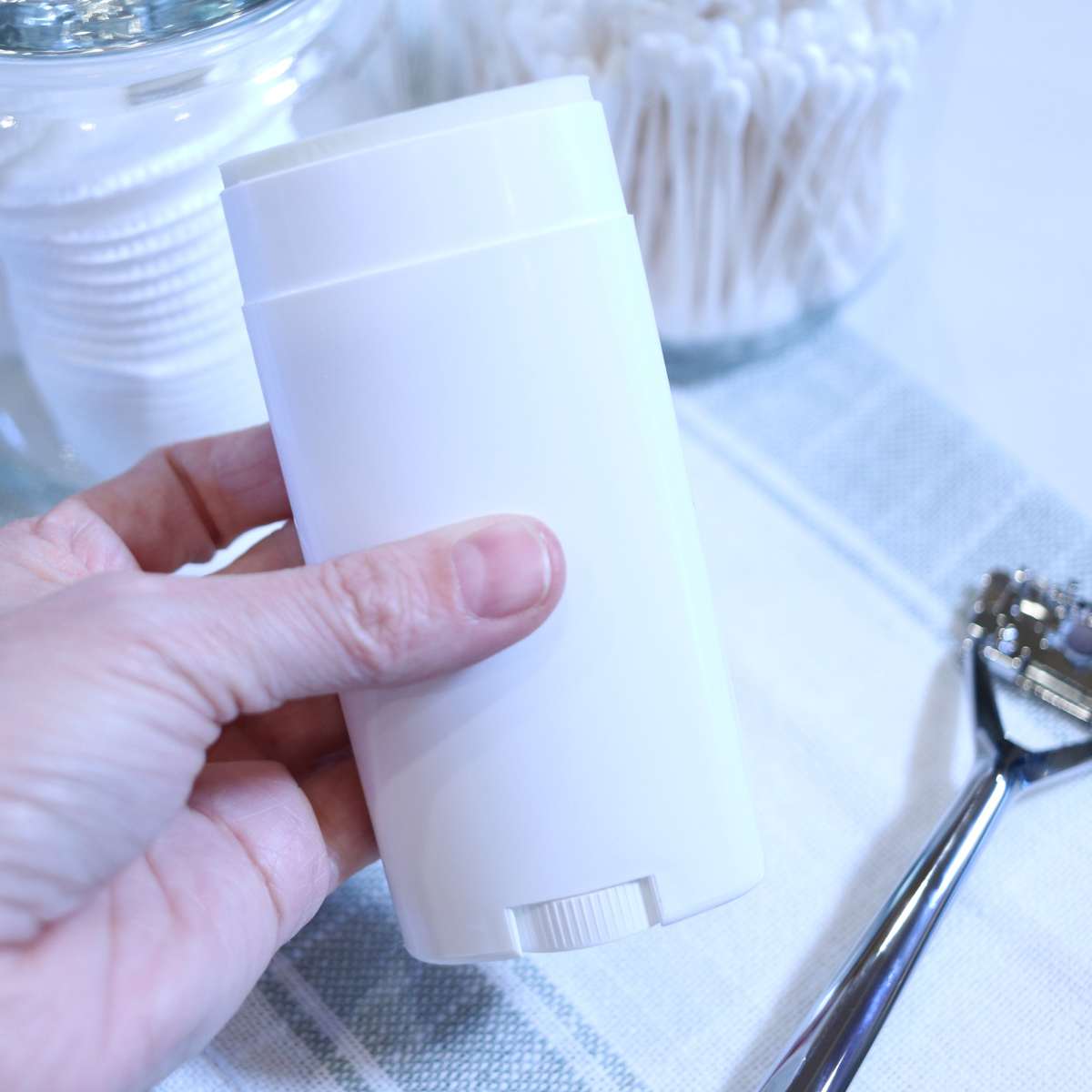 A womans hand is holding onto a white plastic tube of DIY natural deodorant she just made. A silver razor is also sitting on the table near glass jars filled with q-tips and cotton rounds.