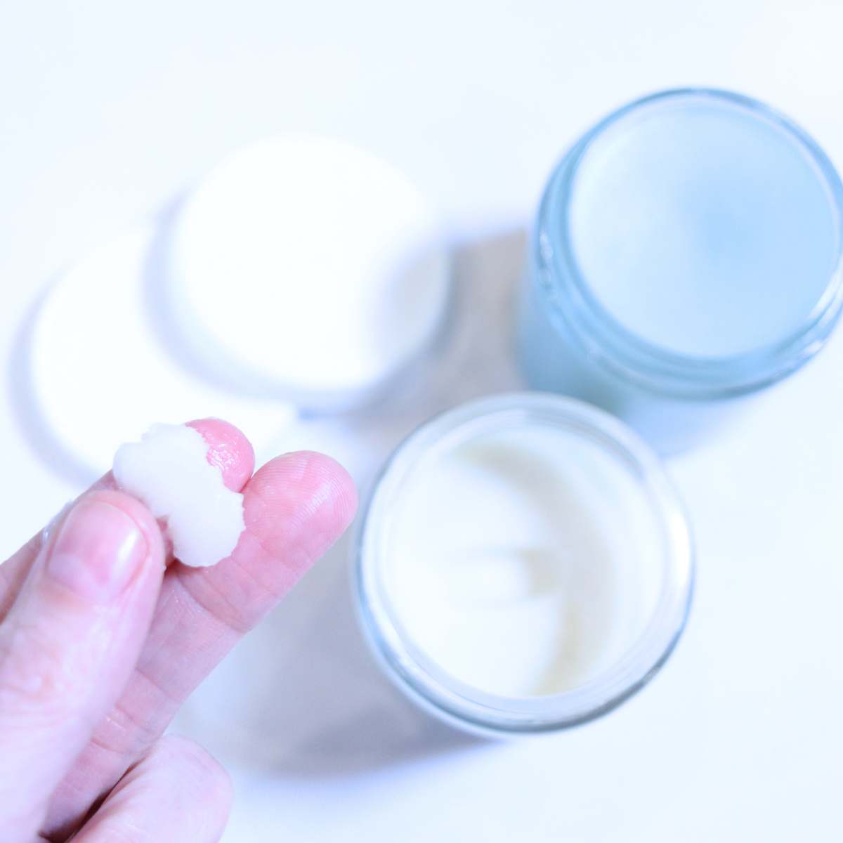 A womans hand is holding up a pea sized amount of DIY natural deodorant with essential oils. two smaller jars of homemade deodorant are in the background.