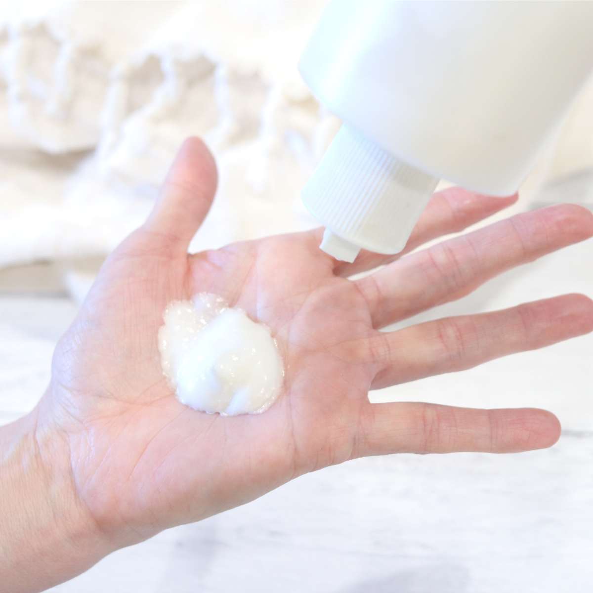 A woman's hand with some white homemade sunscreen on her palm. she is holding a bottle of the sunscreen with the other hand. A cream beach towel is in the background.