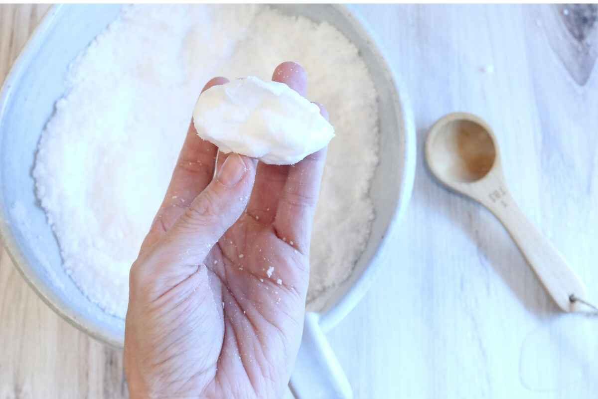 A womans hand is hold a white clump of baking soda and citric acid. She is checking to see if the consistency is right for the homemade toilet bowl cleaner tablets. The mixture is clumping which means it's time to move onto the next step. 