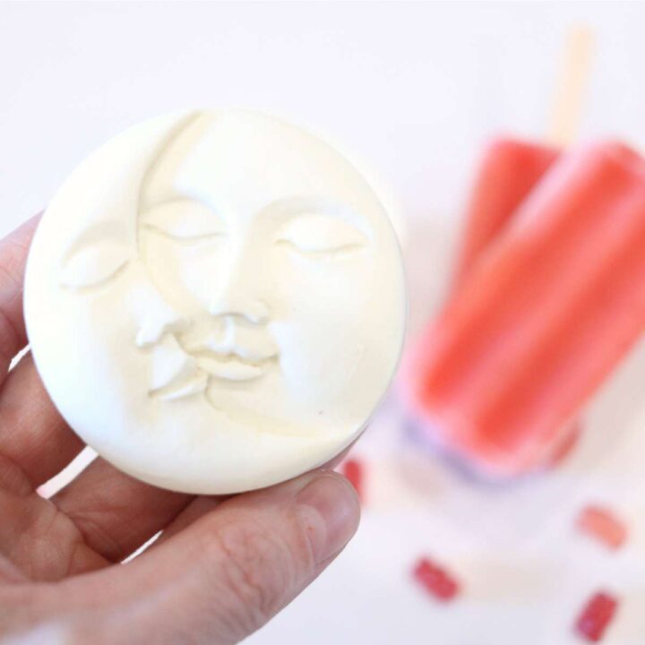 A womans hand is holding a white DIY sunscreen lotion bar to the camera. The bar has an impression of a sun and moon on the front of it. Two pink strawberry popsicles are in the background of the photgraph.
