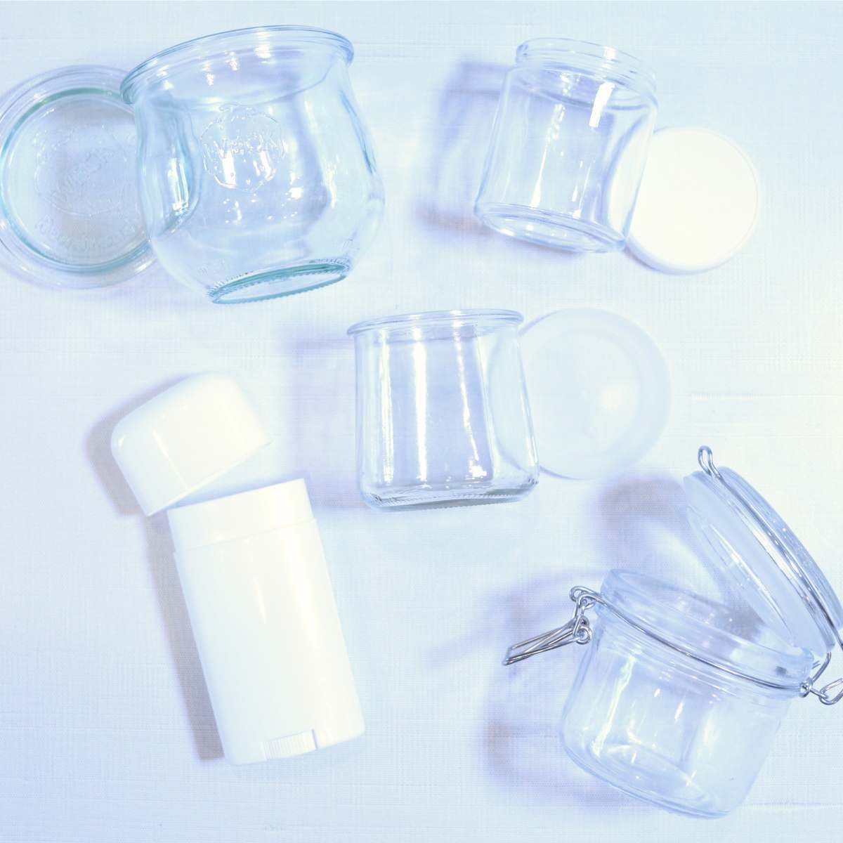 Five different storage containers for DIY deodorant. Four containers are clear glass with plastic lids and one is a white plastic deodorant container.