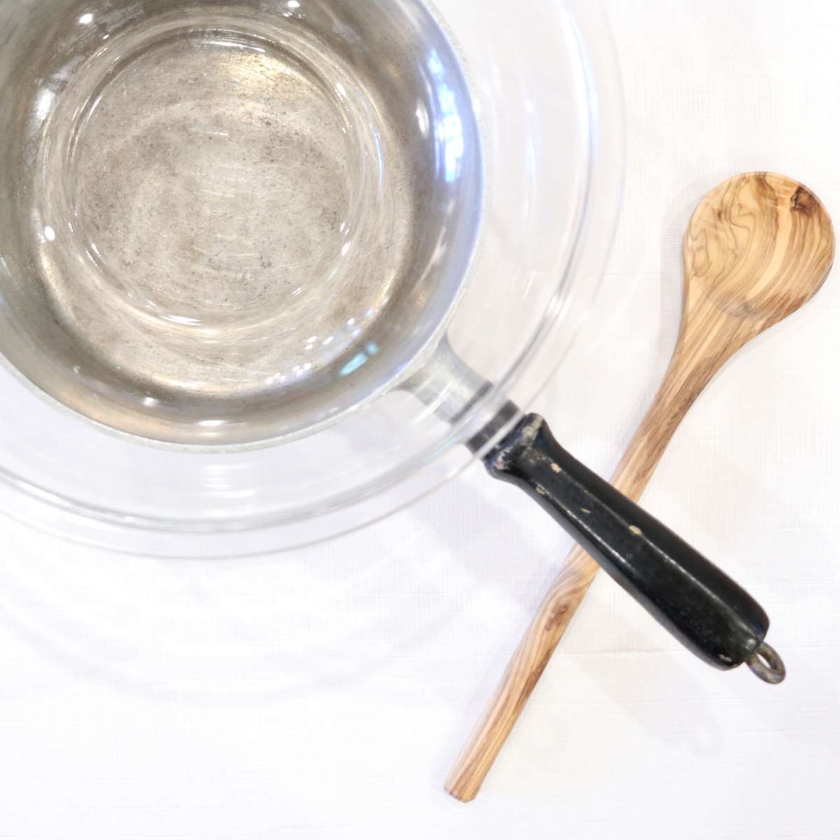 A DIY double broiler using a silver thrifted pot and a clear glass bowl sitting on top. A wooden spoon is nearby for storing ingredients.