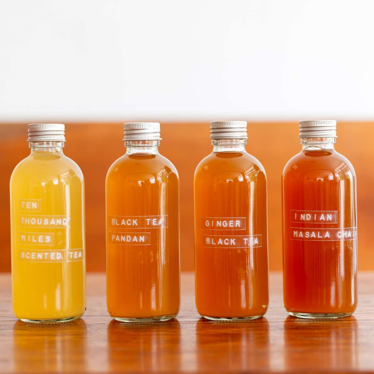 Four bottles of different flavored homemade kombucha lined up on a table. A label is on the front of each bottle indicating the flavor in each. 