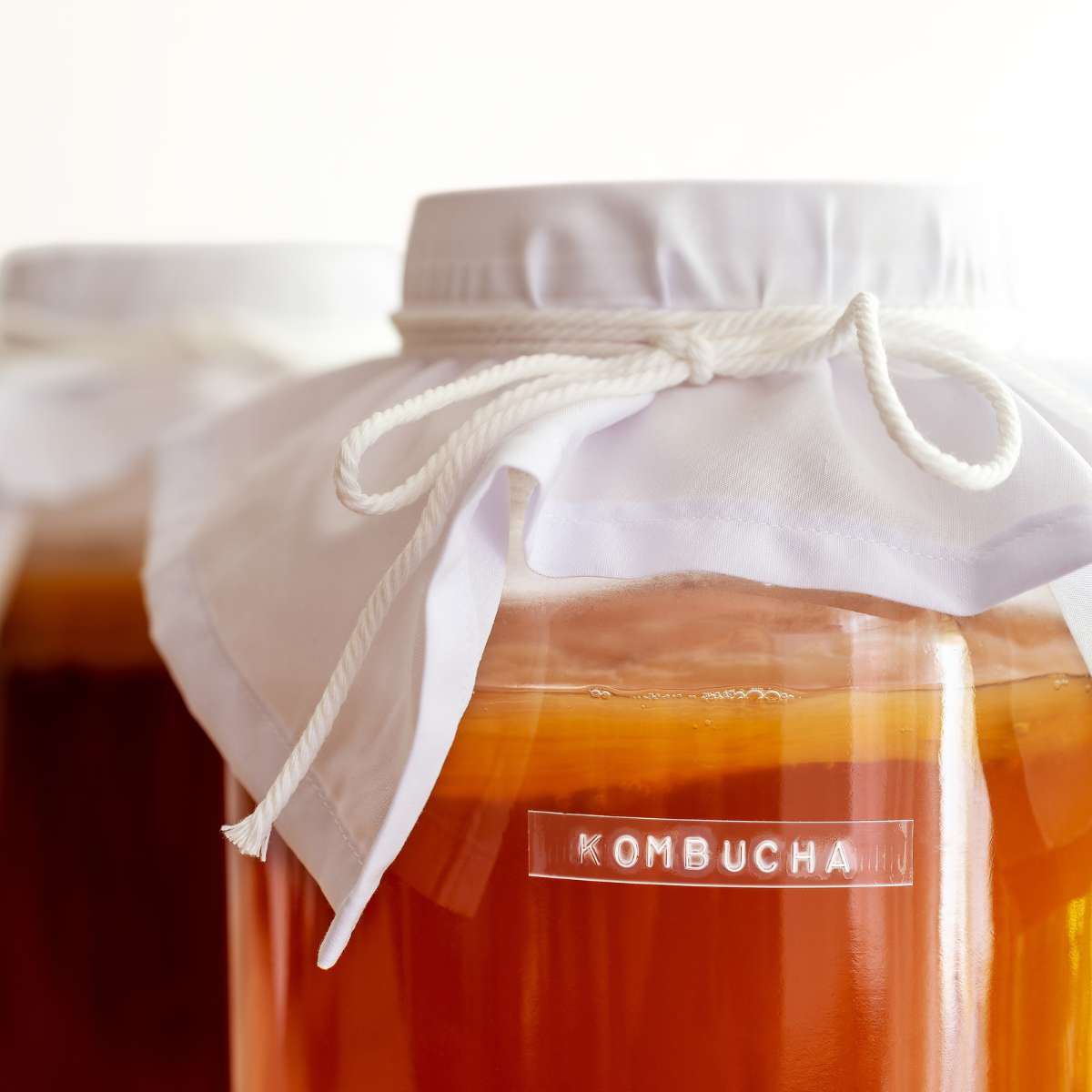 A large clear jar with a label on the front that says kombucha on it. The jar is full of black tea kombucha that has just finished brewing. The white scoby is visible on the top of the kombucha. A white towel tied with string is covering the top of the jar. Another jar of kombucha sits in the background.