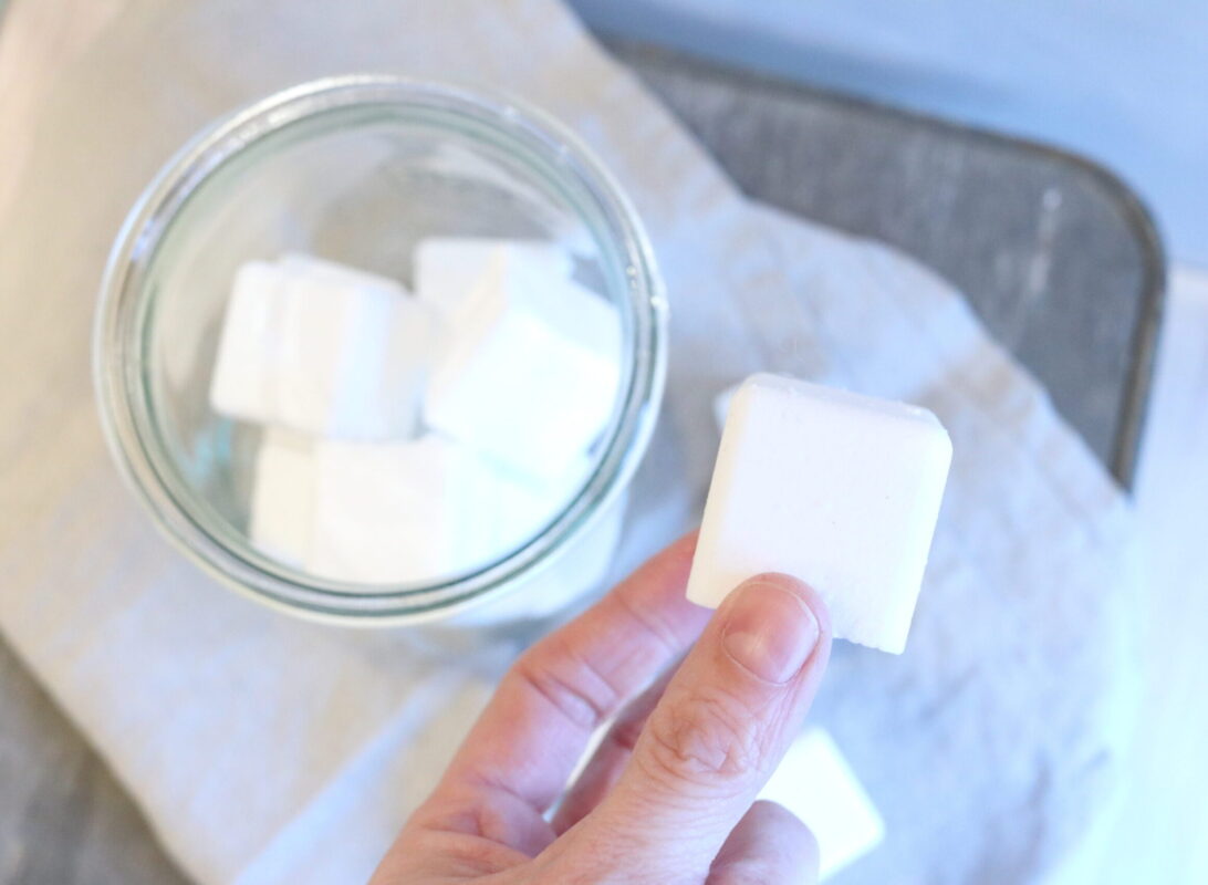 A hand is holding onto a white square cube. The cube is a DIY toilet bowl tablet that was made of simple household ingredients.