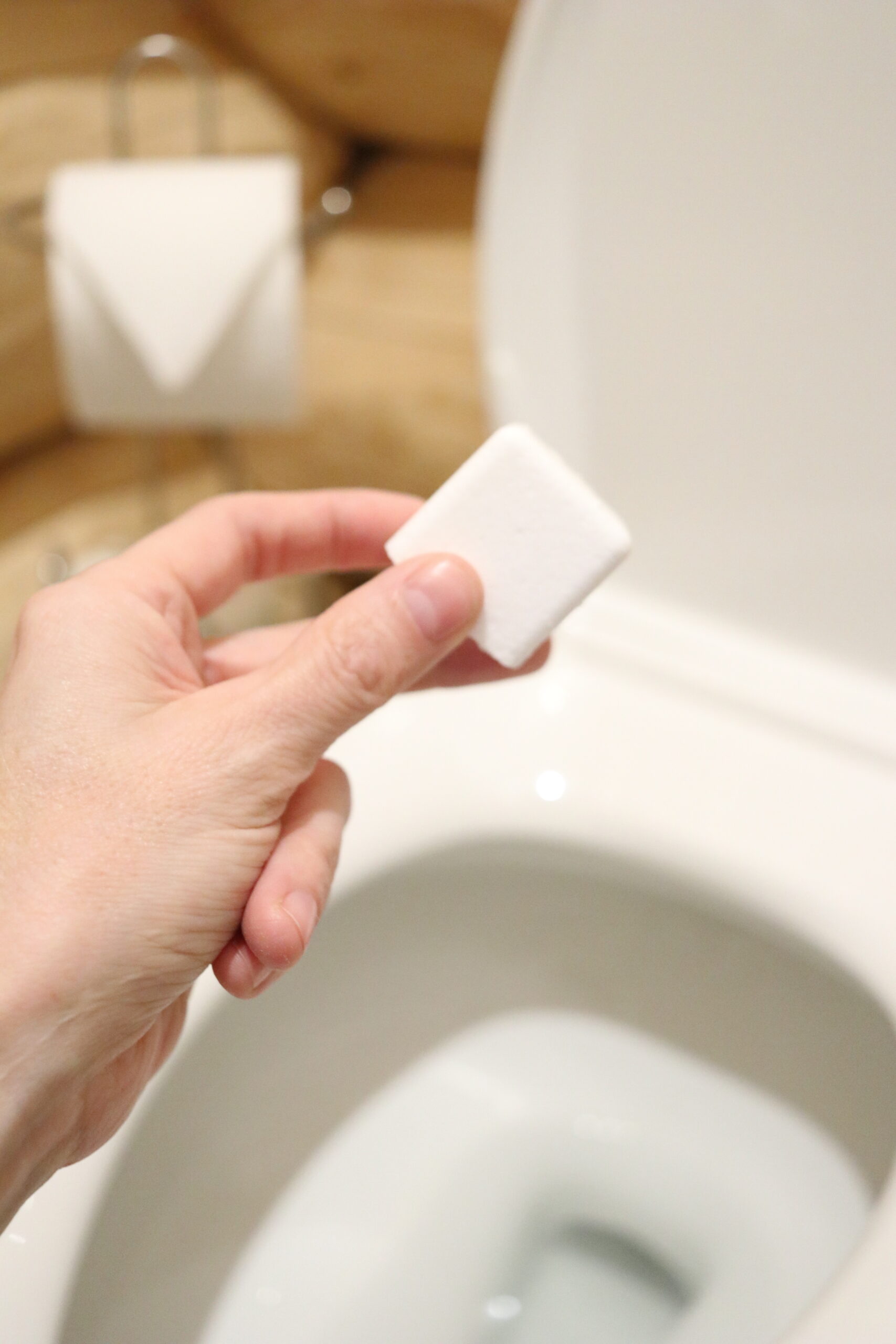 A womans hand is holding a white DIY toilet bowl cleaner tablet. The tablet is made from natural ingredients.