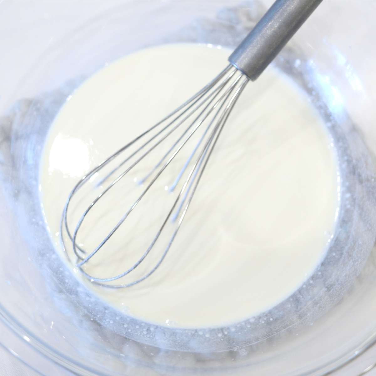 A silver whisk sitting in a clear bowl full of white liquid. The liquid are ingredients being used to make waterproof sunscreen bars that are safe to use on acne prone skin.