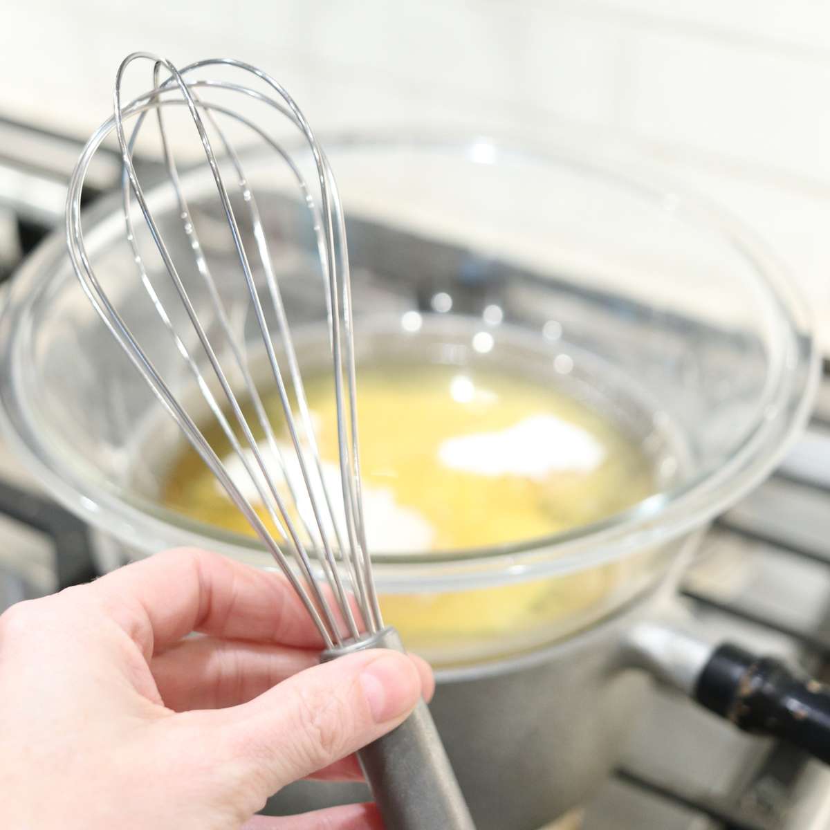 A womans hand is holding a silver metal whisk. A pot behind her is melting ingredients for a homemade natural sunscreen recipe.