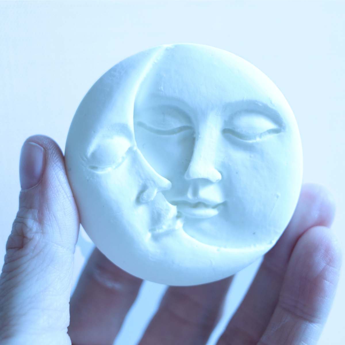 A womans hand holding onto a white sun and moon homemade sunscreen bar in front of a white backdrop. 