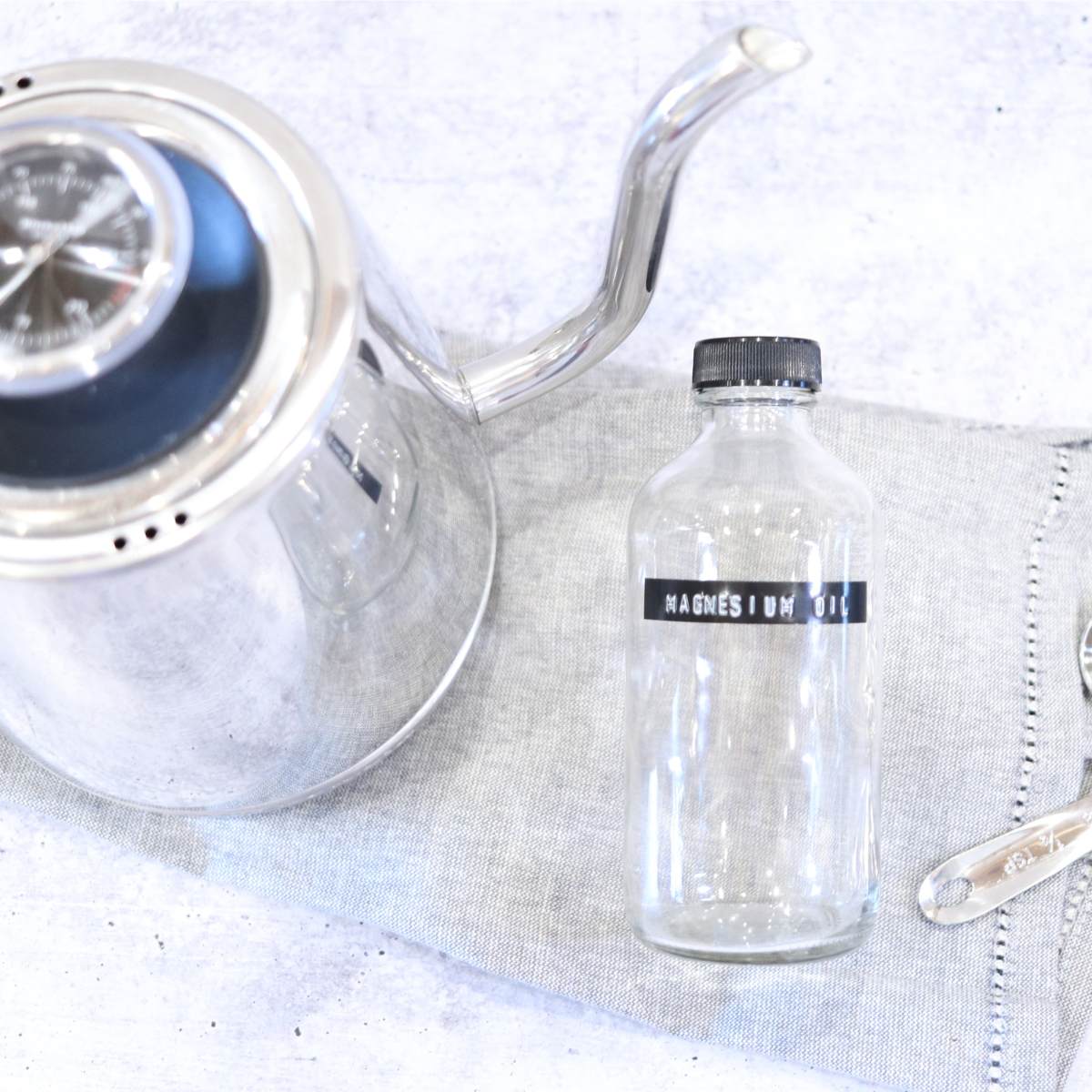 A picture of a shiny chrome electric tea kettle and a clear bottle for magnesium oil resting on a gray linen napkin.