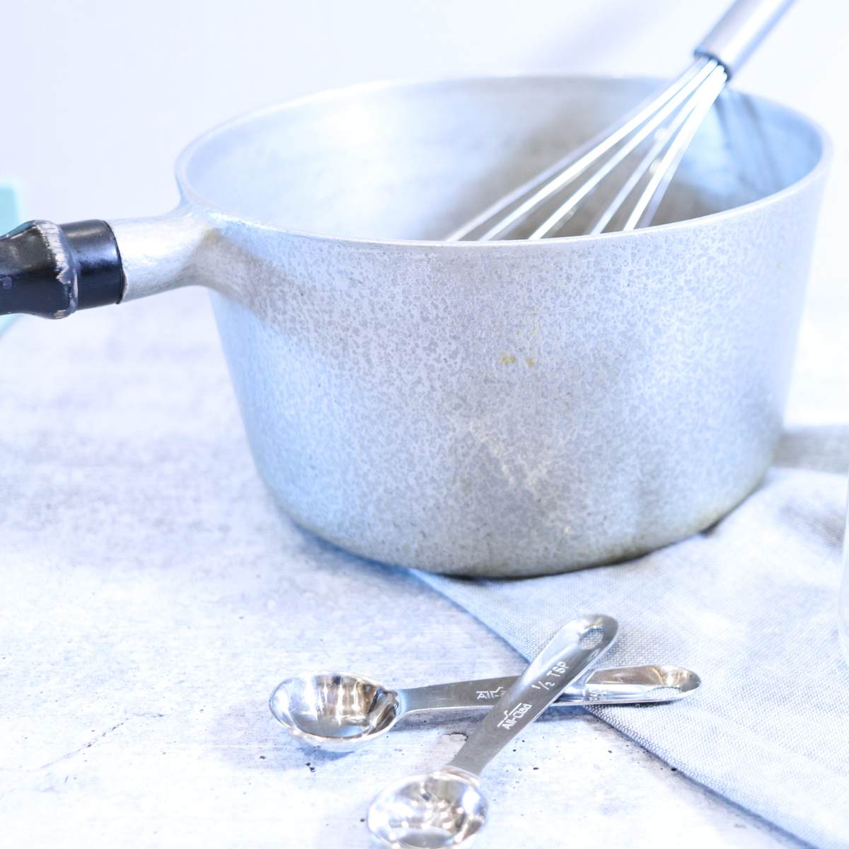 A vintage aluminum pot with a black wooden handle and silver whisk resting ontop of a gray linen napkin. Two silver measuring spoons are resting on the table in front of the pot that will be used for making a recipe.