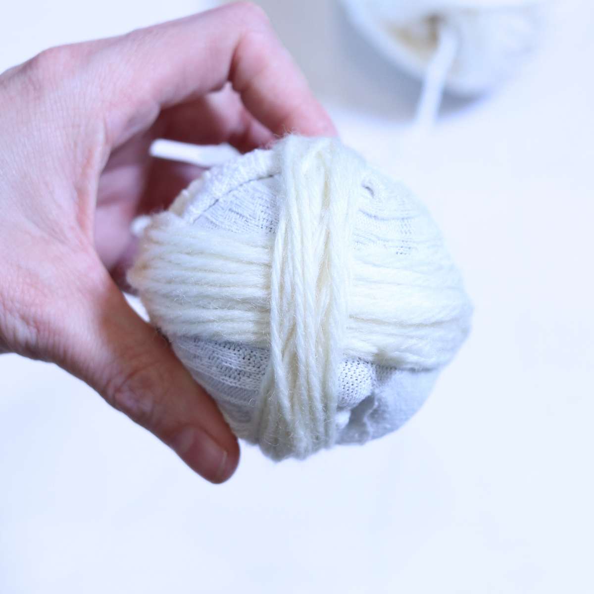 A homemade dryer ball in progress. A womans hand is holding onto the ball that has been wrapped with cream colored yarn.