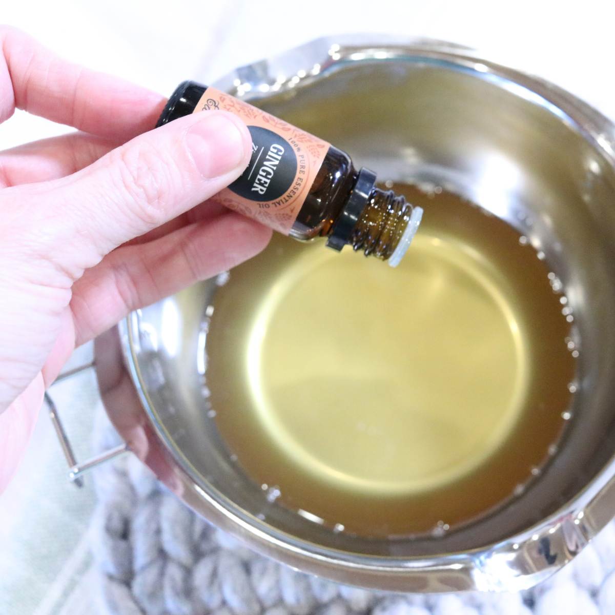 A womans hand is holding an amber bottle of Ginger essential oil over a pot of melted ingredients for a homemade vapor rub with essential oil recipe.