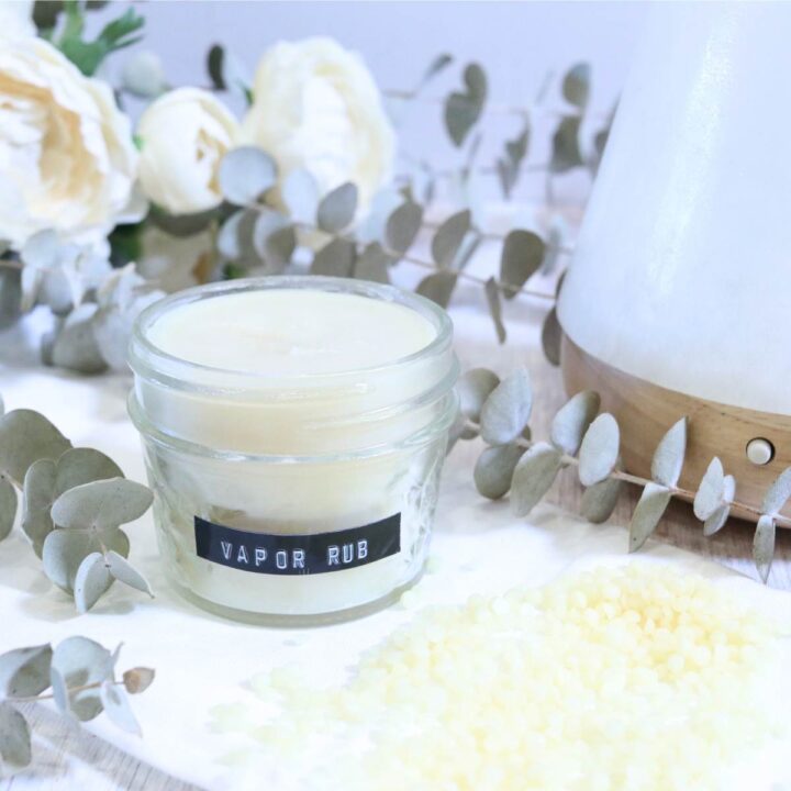 A clear glass jar filled with a pale yellow all-natural vapor rub with soothing essential oils. Some eucalyptus stems and a stone diffuser with a pale wooden base are sitting in the background. A small pile of yellow beeswax pellets sits next to the jar.