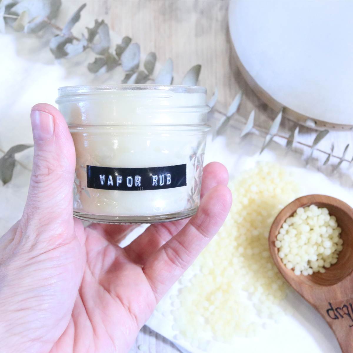 A womans hand is gently holding a clear glass jar with a black and white label that reads vapor rub on it. A stem of greens can be seen in the background of the photo as well as a white topped stone diffuser. A wooden measuring spoon holds a small amount of beeswax pellets.