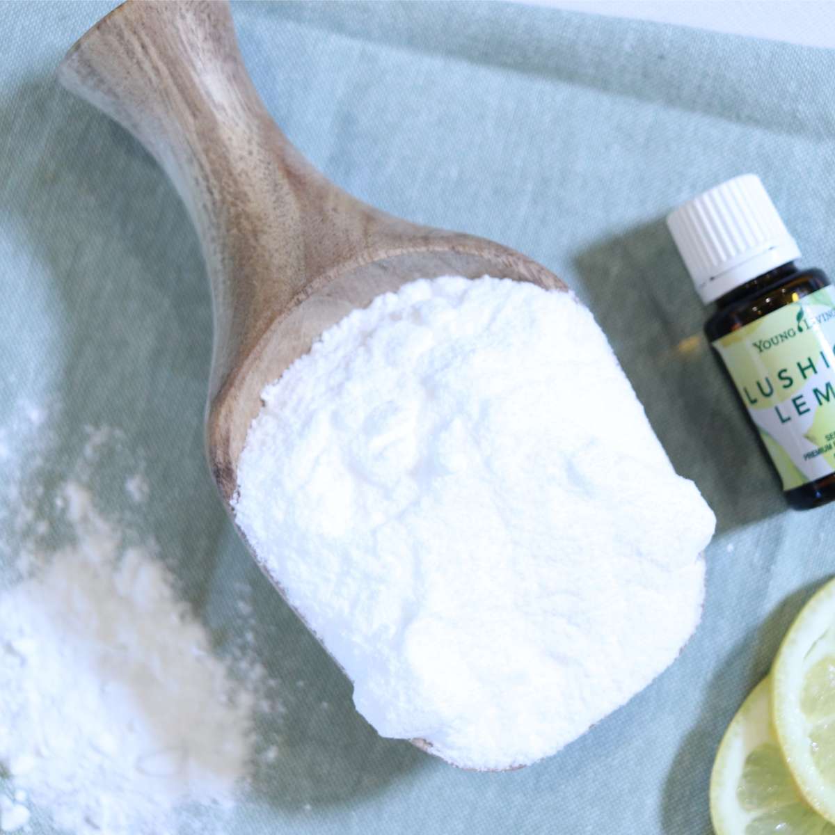 Baking soda based DIY laundry scent booster recipe. A bottle of lemon essential oil and lemon slices are near a wooden scoop filled with baking soda for this scent booster alternative.